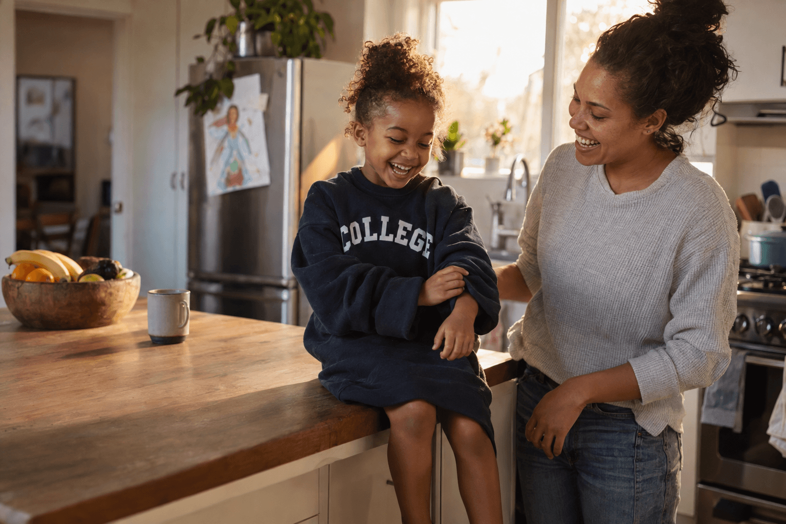 A young girl laughs in an oversized college sweatshirt while her mother smiles beside her in their kitchen.